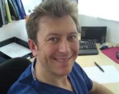 Glyn Thomas, man wearing blue scrubs sitting and desk and smiling
